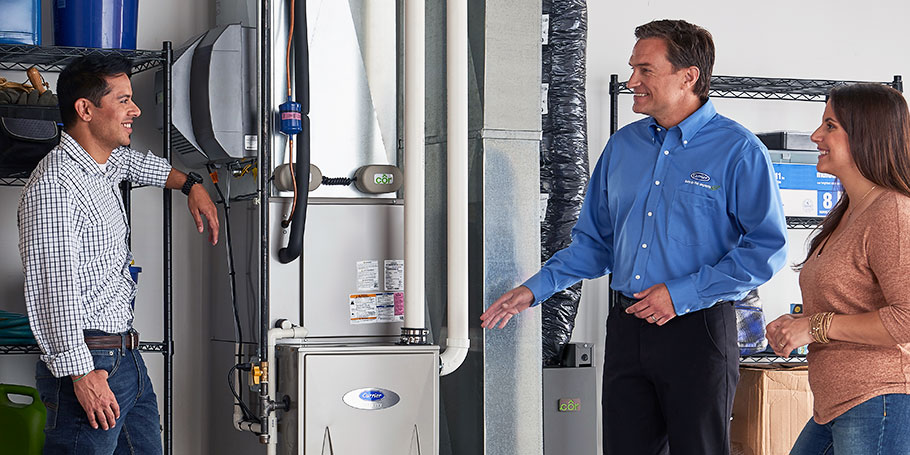 Three people stand in a garage discussing a heating unit. One man gestures toward the unit, while the others listen. Shelving with various items is visible in the background.