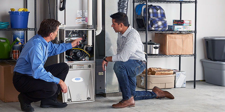 A technician and a man examine a home furnace in a garage with shelves holding household items.