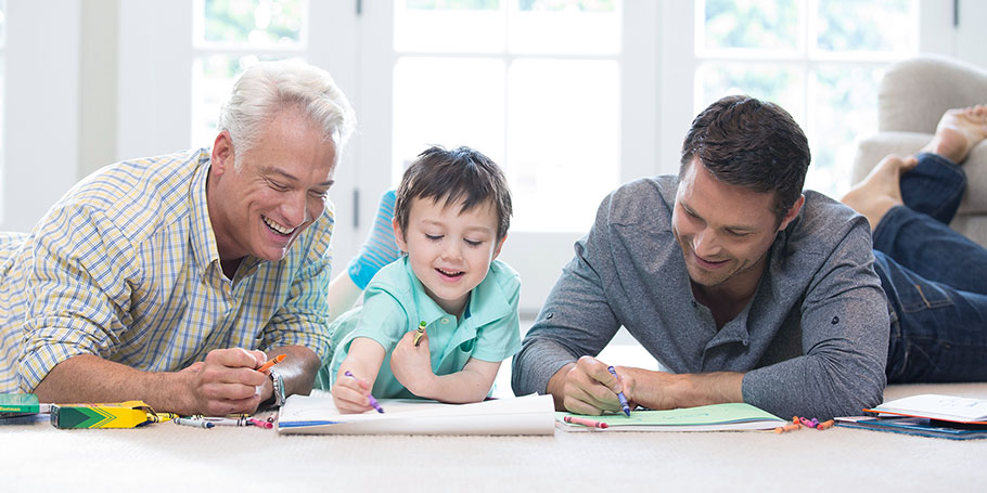Two adults and a child lying on the floor, drawing with crayons on paper. The room is bright with natural light from a window.