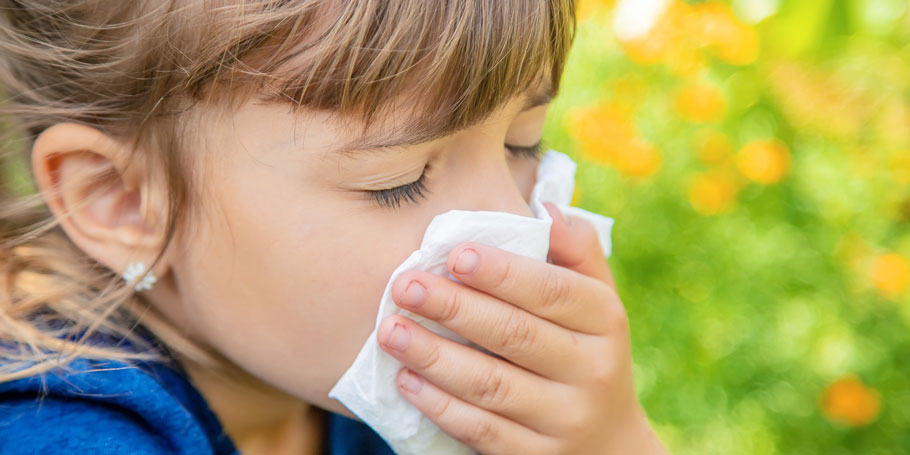 Child with closed eyes holds a tissue to their nose, appearing to sneeze or blow their nose, against a blurred outdoor background.