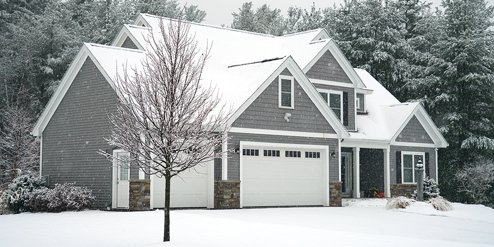 Two-story gray house with snow-covered roof and yard, surrounded by snow-laden trees. A bare tree stands in the foreground.