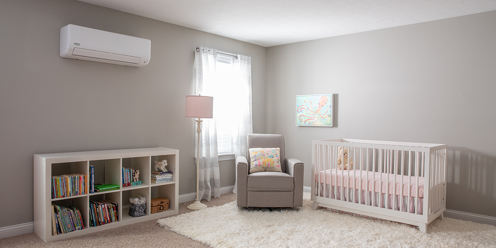 A nursery with a crib, bookshelf, armchair, and lamp. Walls are gray, and there's a plush rug on the floor. A split air conditioner is mounted on the wall.