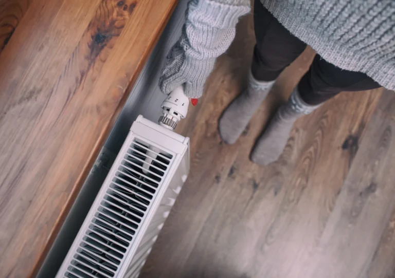 %Imagetitle% | New Berlin Heating &Amp; Air Conditioning Person in a gray sweater and gloves adjusting a thermostat on a white radiator, standing on wooden flooring.