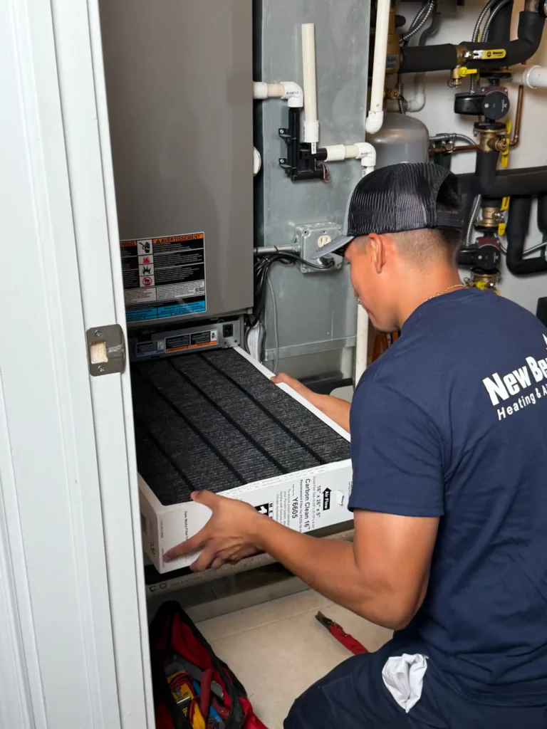 A technician wearing a cap replaces an air filter in an HVAC system inside a utility closet.
