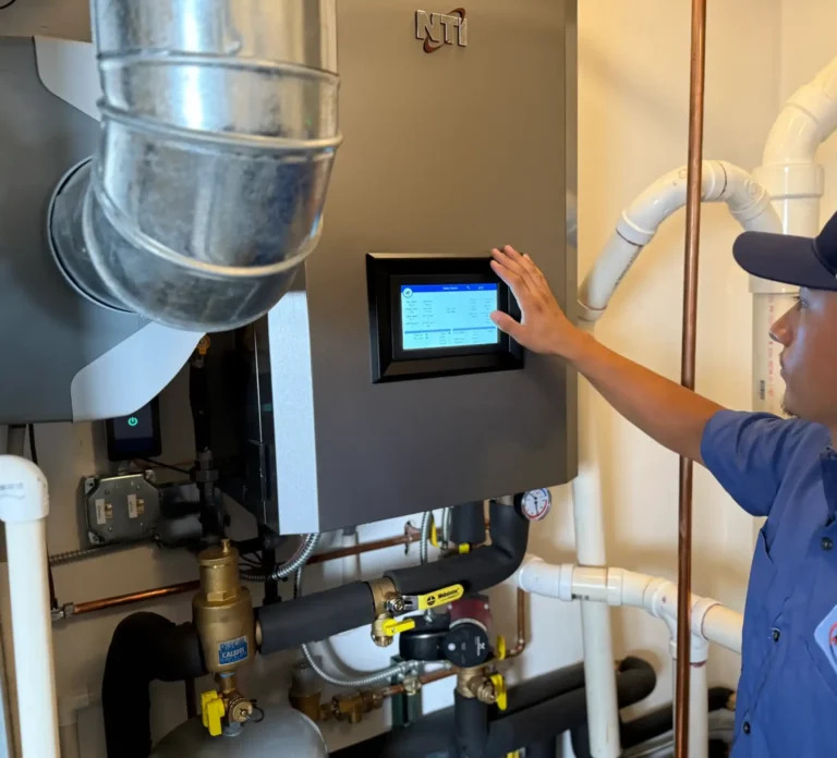 A technician in uniform operates a touchscreen control panel on a heating or HVAC system surrounded by pipes and ducts.