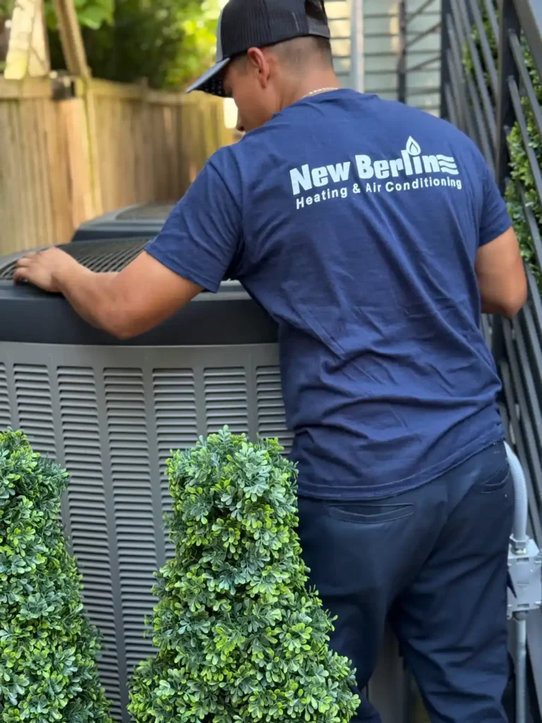 A technician wearing a "New Berlin Heating & Air Conditioning" shirt services a large outdoor HVAC unit beside a fence and artificial bushes.
