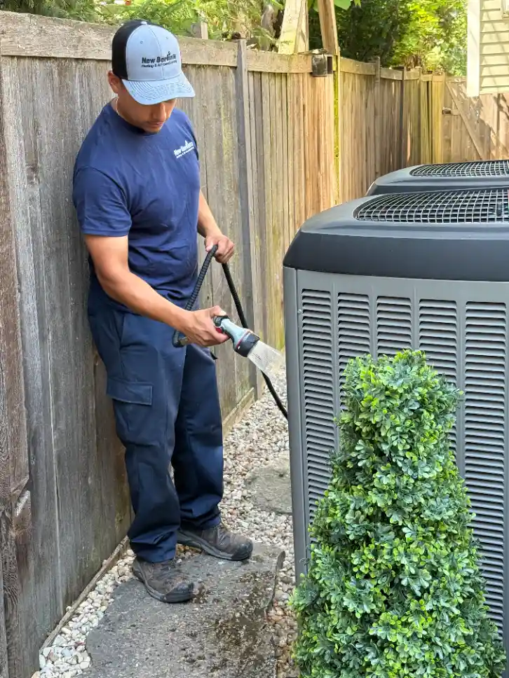 A man in work clothes uses a hose to clean an outdoor air conditioning unit beside a wooden fence and a small shrub.