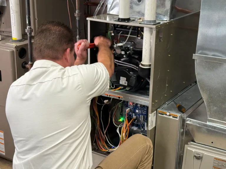 A technician kneels and works on the internal wiring of an open HVAC unit with various visible components and wires.