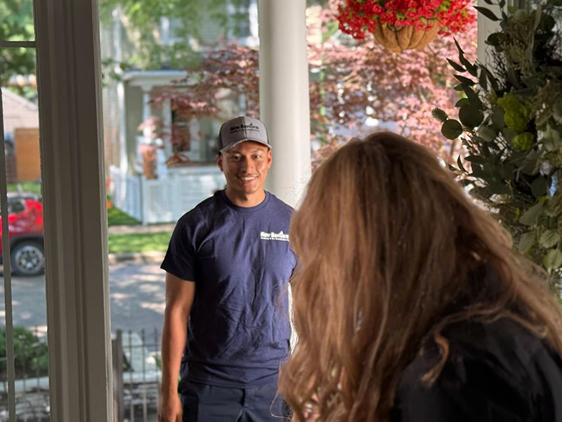 A delivery worker in a blue shirt and cap stands outside a front door, smiling at a person with long hair inside the house.