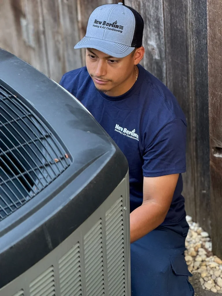 A technician wearing a "New Berlin Heating & Air Conditioning" shirt and hat works on an outdoor AC unit next to a wooden fence.