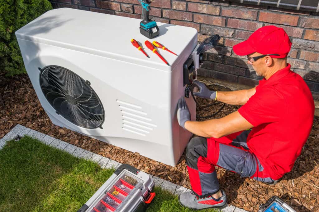 Technician in red uniform servicing an outdoor heat pump unit, using tools placed on top of the unit, with a toolbox and drill nearby on the ground—a typical scene when ensuring that heat pumps work efficiently, even in tough Wisconsin winters.