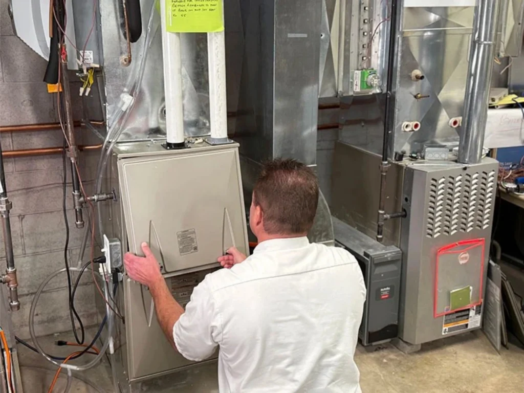 A technician in a white shirt works on the front panel of a furnace unit in a mechanical room with various HVAC equipment.